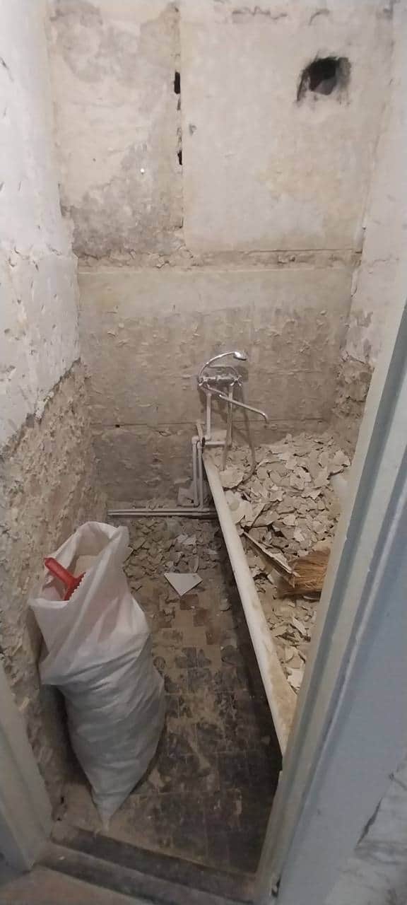 Baie de bloc în demolare: faianța veche desprinsă, moloz pe pardoseală și sac de resturi lângă bateria demontată.